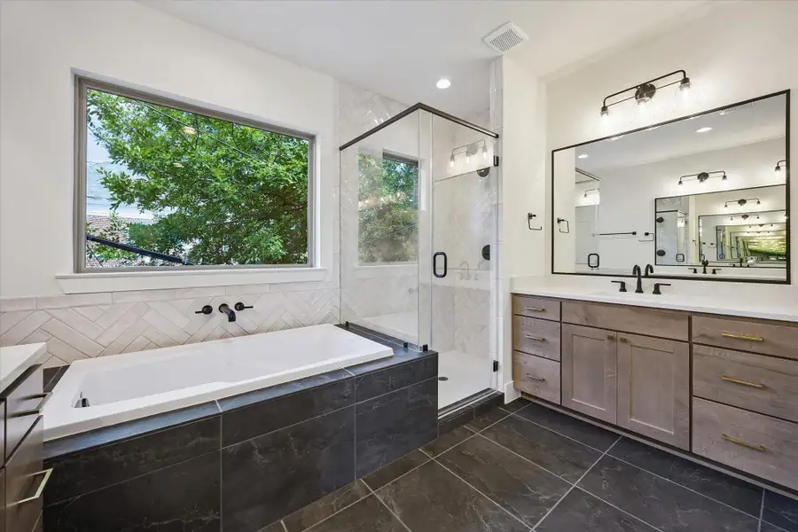 The luxurious deep soaking tub is framed by lush greenery. The space is accented by a sleek wall-mounted spout and a stunning glass-enclosed shower, beautifully finished with intricate, floor-to-ceiling herringbone tile.