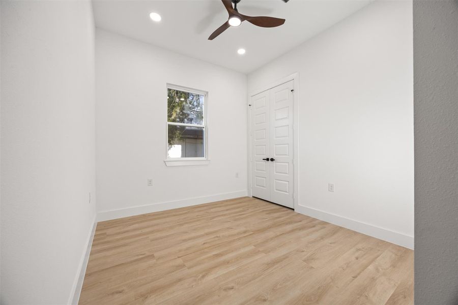 Bright, minimalist room with wood-like flooring, ceiling fan, single window, and double closet doors. Ideal for a small bedroom or office.