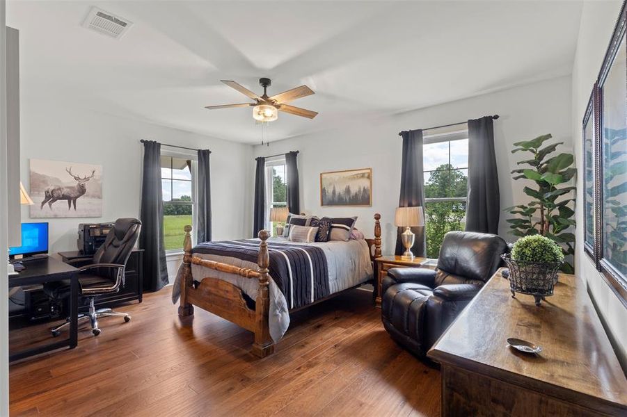Spacious primary bedroom suite featuring rich hardwood flooring, multiple windows with serene acreage views, and room for a sitting area or private workspace—blending comfort, function, and refined country living.
