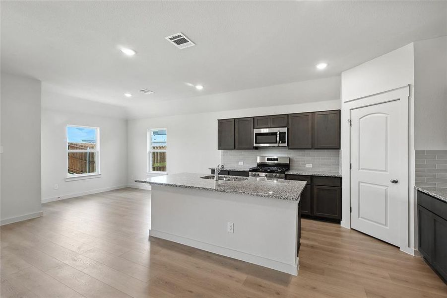 Kitchen featuring light stone countertops, stainless steel appliances, a center island with sink, dark wood finish cabinets, and light wood-style floors