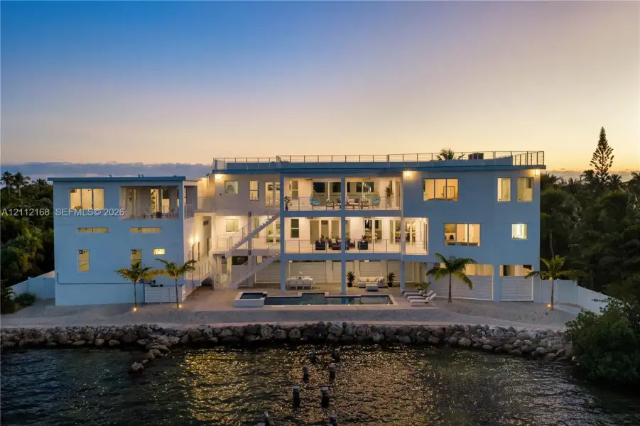 Waterfront multi-story residence featuring a private swimming pool, expansive stacked balconies with metal railings, a rooftop terrace, and a stone-lined bulkhead Waterfront multi-story residence featuring a private swimming pool, expansive stacked balconies with metal railings, a rooftop terrace, and a stone-lined bulkhead