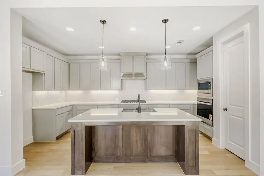 Two tone kitchen featuring a kitchen island with sink, light wood-style floors, decorative backsplash, pendant lighting, and two tone color scheme
