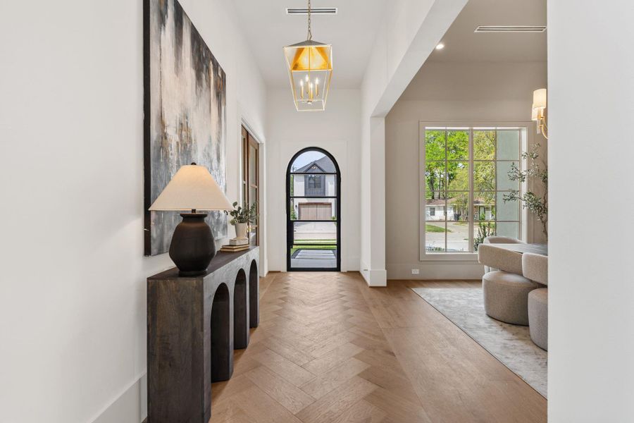 A welcoming entry unfolds with soaring ceilings and wide-plank herringbone hardwood floors, leading the eye toward an arched glass front door. Natural light pours in through expansive front-facing windows, while a statement pendant adds warmth and dimension to the space.