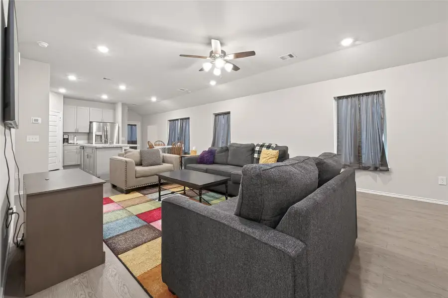 This open-concept living space features a modern kitchen with stainless steel appliances, a comfortable seating area with a large sectional sofa, and a colorful area rug. The room is well-lit with recessed lighting and a ceiling fan, creating a bright and inviting atmosphere.