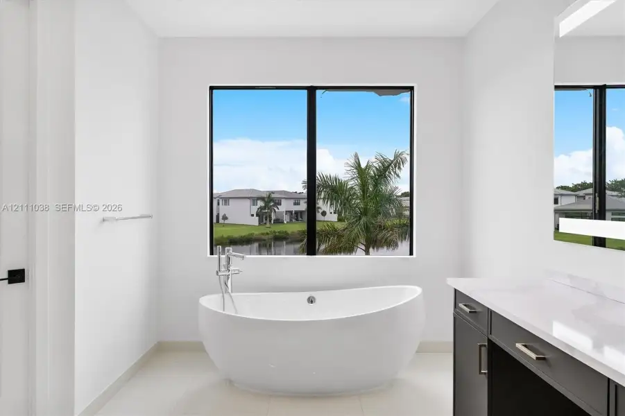 Freestanding soaking tub with chrome floor-mounted faucet, dark cabinetry featuring rectangular hardware, solid-surface countertop, and back-lit mirror