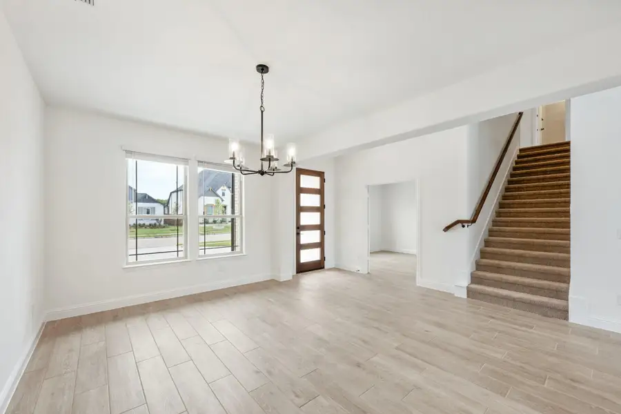 Open living area with light wood floors, chandelier, wood front door, and carpeted staircase nearby
