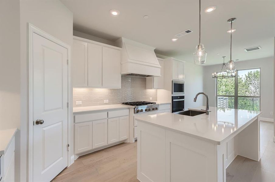 Kitchen with light wood-type flooring, custom range hood, appliances with stainless steel finishes, backsplash, and a kitchen island with sink