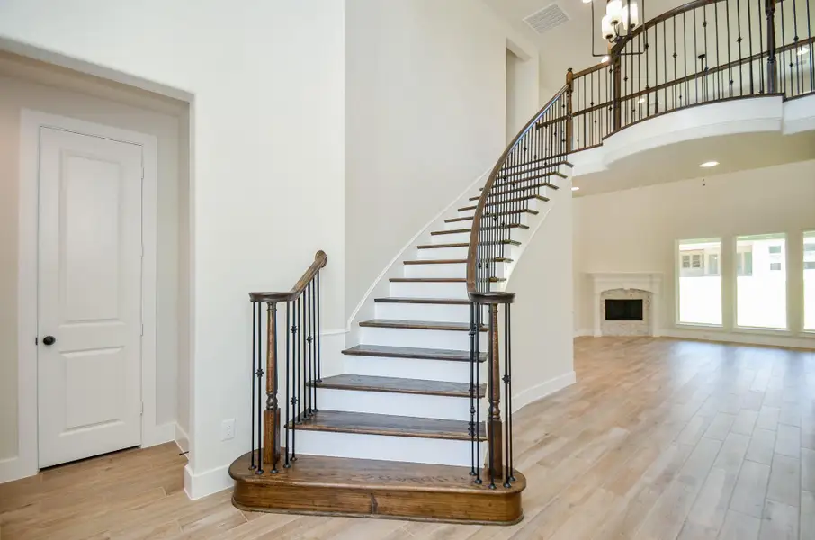 This image showcases a stunning curved staircase with dark wood steps and wrought iron balusters. The staircase leads up to a balcony overlooking the open-concept living space. The room features high ceilings, wide plank flooring, and a beautifully framed fireplace in the corner with large windows that offer plenty of natural light.