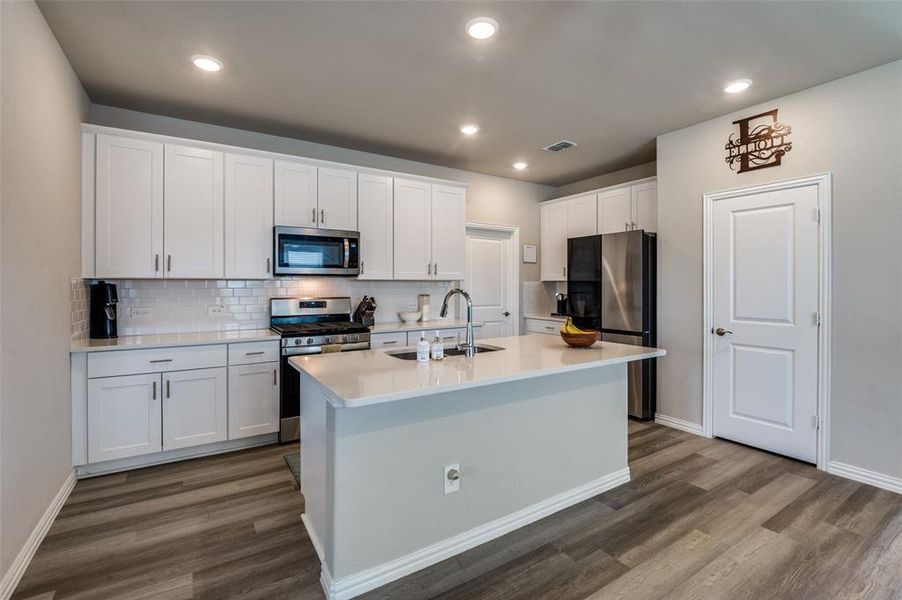 Kitchen featuring white cabinets, stainless steel appliances, a center island with sink, dark wood-style flooring, and tasteful backsplash