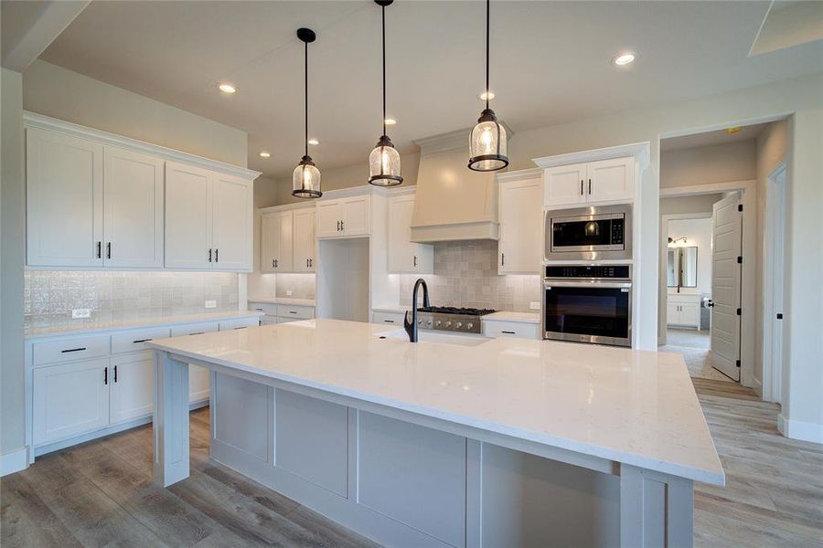 Kitchen featuring light wood-type flooring, a large island with sink, backsplash, white cabinets, and appliances with stainless steel finishes