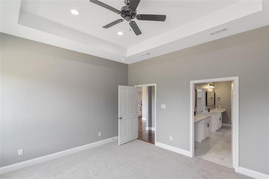 Unfurnished bedroom featuring light colored carpet, ensuite bathroom, a tray ceiling, and ceiling fan