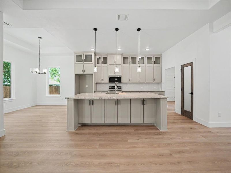 Open-concept kitchen featuring a large central island with a light-toned countertop, grey shaker-style cabinetry, and three pendant lights Open-concept kitchen featuring a large central island with a light-toned countertop, grey shaker-style cabinetry, and three pendant lights