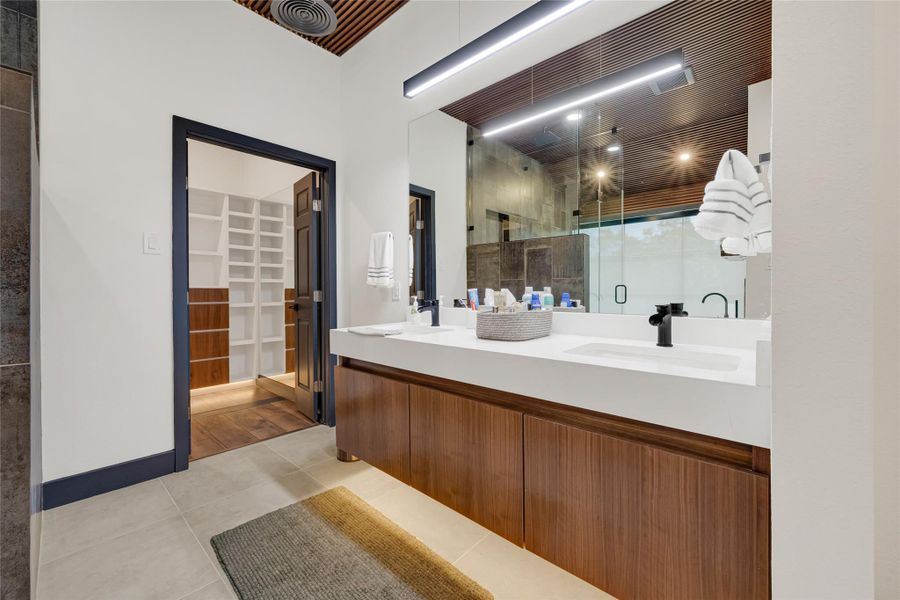 Double vanity with ample storage, a large mirror, and stylish lighting. The room is connected to a spacious walk-in closet with built-in shelving, and the shower area is enclosed with glass, enhancing the contemporary design.