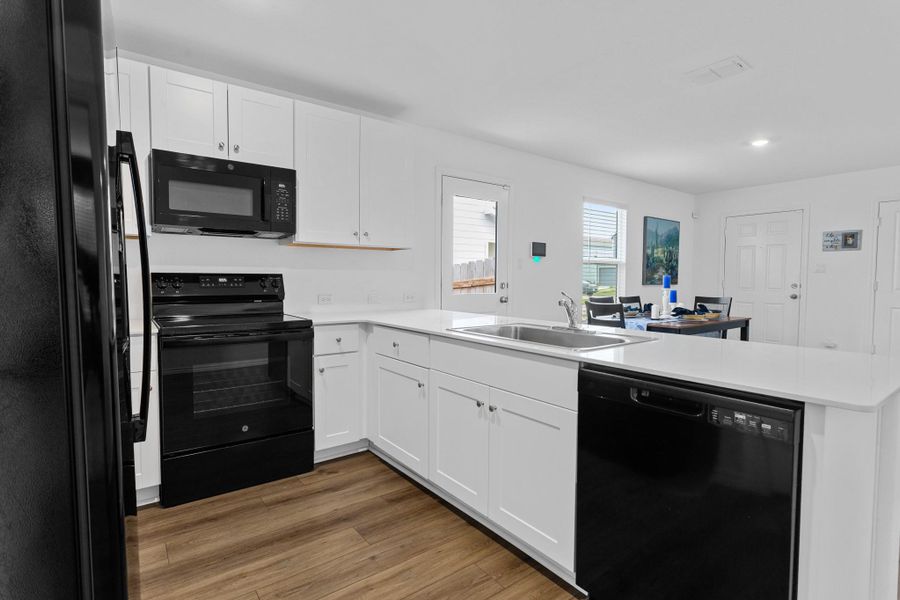 Open-concept kitchen featuring white shaker cabinetry, an integrated stainless steel sink, black appliance suite, white countertops, and wood-finish flooring