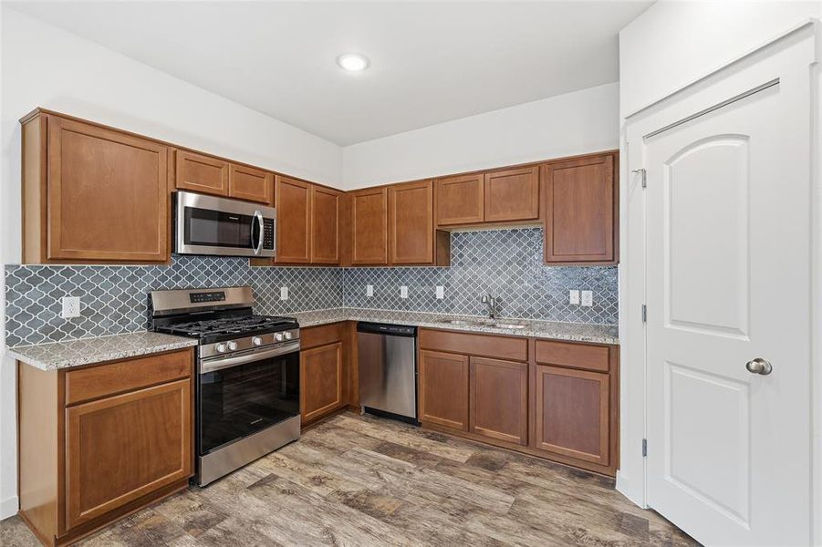 Kitchen with stainless steel appliances, brown cabinetry, light stone countertops, and decorative backsplash Kitchen with stainless steel appliances, brown cabinetry, light stone countertops, and decorative backsplash