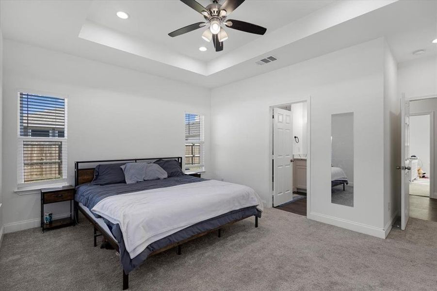 Carpeted bedroom featuring a raised ceiling, multiple windows, ceiling fan, recessed lighting, and connected bathroom Carpeted bedroom featuring a raised ceiling, multiple windows, ceiling fan, recessed lighting, and connected bathroom