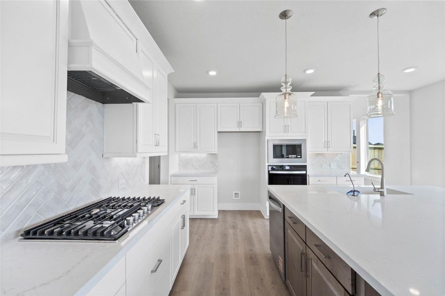 The open-concept gourmet kitchen designed for both cooking and entertaining. It features elegant white shaker-style cabinetry, pristine quartz countertops, and a large central island that doubles as a functional breakfast bar. The cooking area is a chef's dream with a sleek stainless steel gas cooktop, hood vent, and oven, all complemented by a chic white tile backsplash in a herringbone pattern.