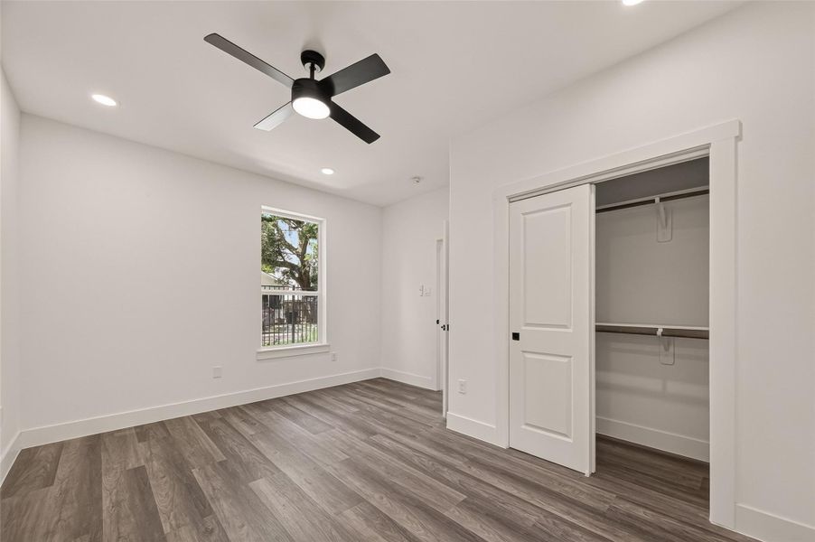 This bright, modern bedroom features sleek wood flooring, a ceiling fan, recessed lighting, and a spacious closet with sliding doors. A large window offers natural light and a view of the outdoors. This bright, modern bedroom features sleek wood flooring, a ceiling fan, recessed lighting, and a spacious closet with sliding doors. A large window offers natural light and a view of the outdoors.