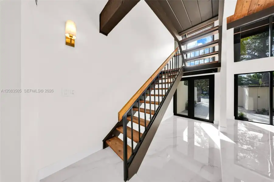 Staircase featuring open wood treads, black metal railings, and a wood handrail, polished white tile flooring, exposed dark wood ceiling beams, and floor-to-ceiling glass paneling Staircase featuring open wood treads, black metal railings, and a wood handrail, polished white tile flooring, exposed dark wood ceiling beams, and floor-to-ceiling glass paneling