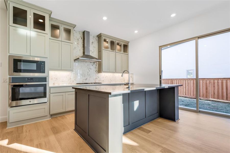 Kitchen featuring built in microwave, wall chimney exhaust hood, stainless steel oven, light wood-type flooring, and gas cooktop