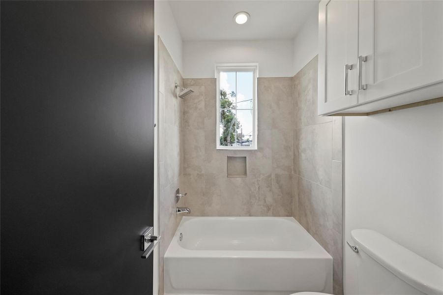Modern bathroom featuring a bathtub/shower combo, elegant neutral tiles, abundant natural light from the window, and built-in shelving for convenient storage. Modern bathroom featuring a bathtub/shower combo, elegant neutral tiles, abundant natural light from the window, and built-in shelving for convenient storage.