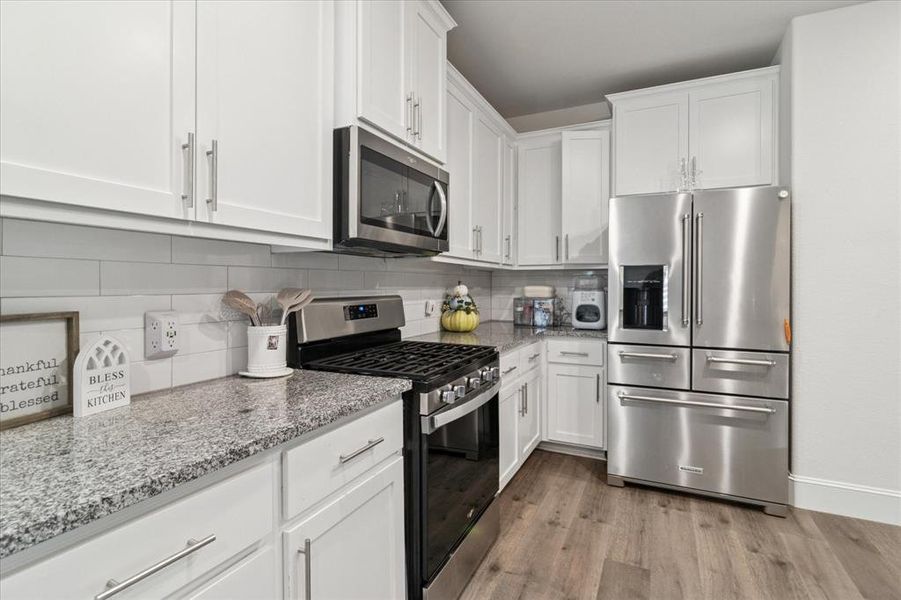 Kitchen featuring appliances with stainless steel finishes, backsplash, light stone countertops, white cabinetry, and light wood-type flooring Kitchen featuring appliances with stainless steel finishes, backsplash, light stone countertops, white cabinetry, and light wood-type flooring