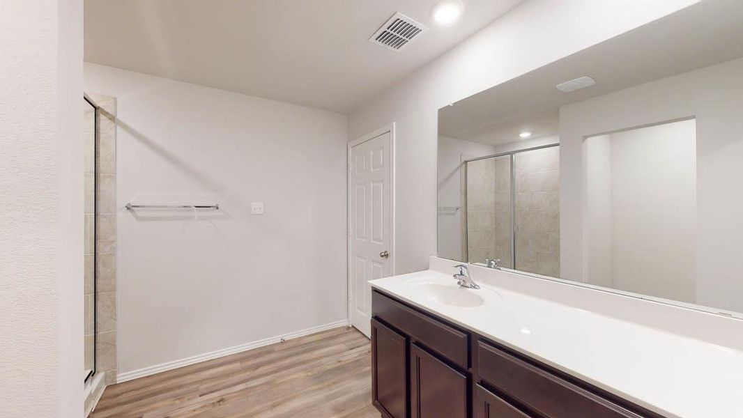 Bathroom with wood-finish flooring, a double vanity featuring a white countertop and dark wood cabinetry, a large wall-mounted mirror, and a glass-enclosed shower with tiled walls