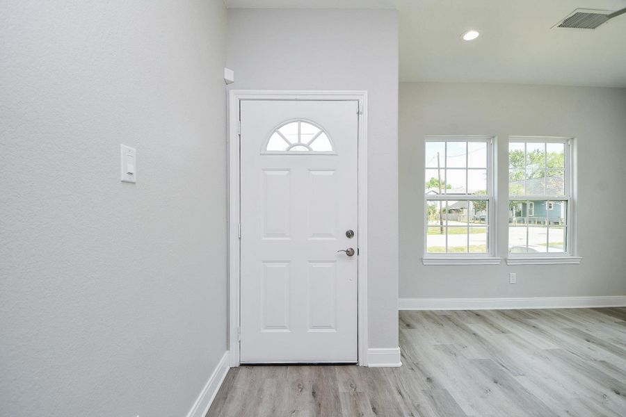 Bright entryway with a modern white front door and two large windows, allowing plenty of natural light. The space features light gray walls and wood-style flooring, creating a fresh and inviting atmosphere.