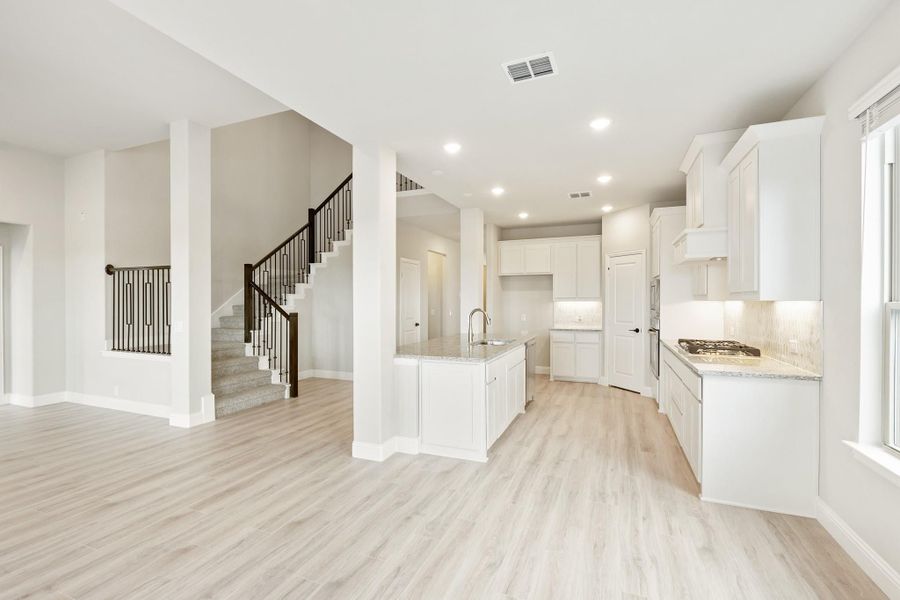 Open-concept kitchen with white cabinets, center island, light wood floors, and staircase visible in background. Open-concept kitchen with white cabinets, center island, light wood floors, and staircase visible in background.