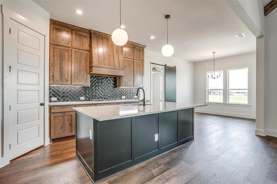 Kitchen with dark hardwood / wood-style flooring, a barn door, custom exhaust hood, decorative light fixtures, and a center island with sink