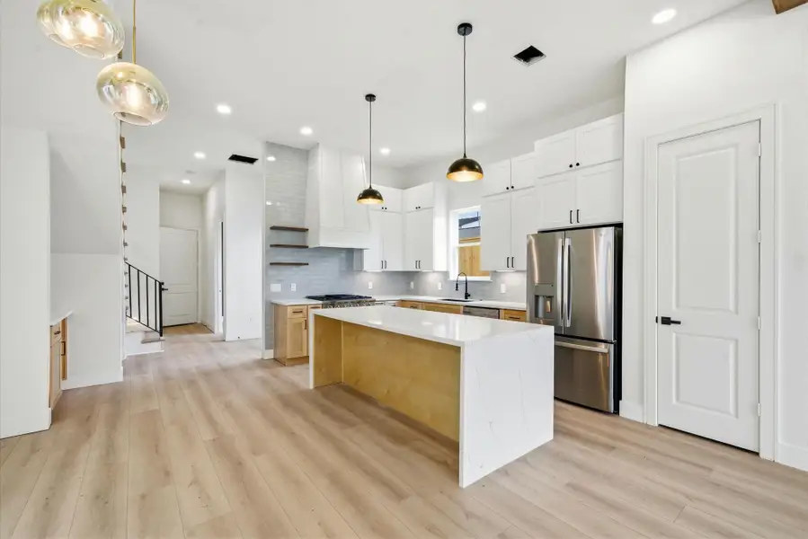 This modern kitchen features sleek white cabinetry, a stylish island with a waterfall countertop, and stainless steel appliances. Pendant lighting and light wood flooring add warmth, while an open layout enhances space and functionality.
