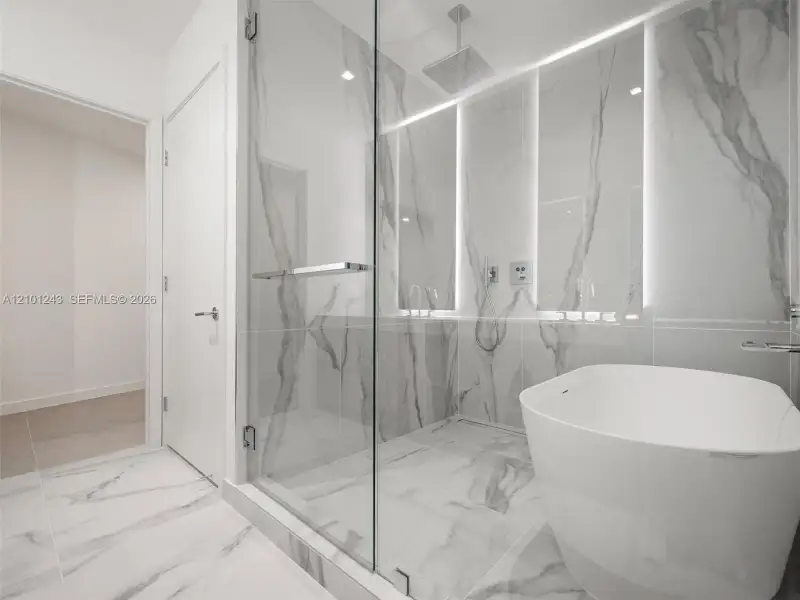 Glass-enclosed shower with large-format veined tile, integrated wall lighting, overhead rain shower head, and freestanding white soaking tub Glass-enclosed shower with large-format veined tile, integrated wall lighting, overhead rain shower head, and freestanding white soaking tub