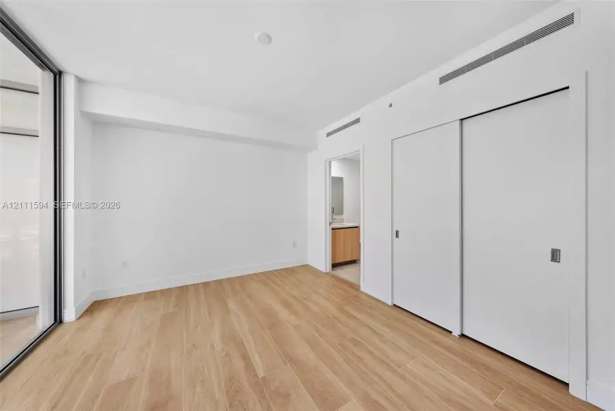 Room featuring wood-finish flooring, full-height sliding door closet, floor-to-ceiling glass wall, and smooth-finish white walls Room featuring wood-finish flooring, full-height sliding door closet, floor-to-ceiling glass wall, and smooth-finish white walls