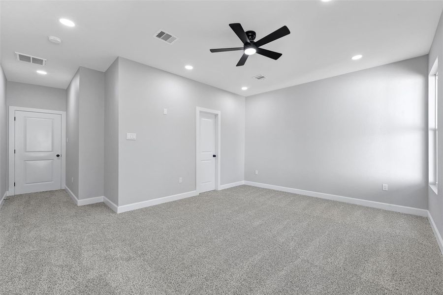 Spacious, light-filled room with neutral gray walls and plush carpet. Features recessed lighting, ceiling fan, and white doors, creating a modern and comfortable ambiance.