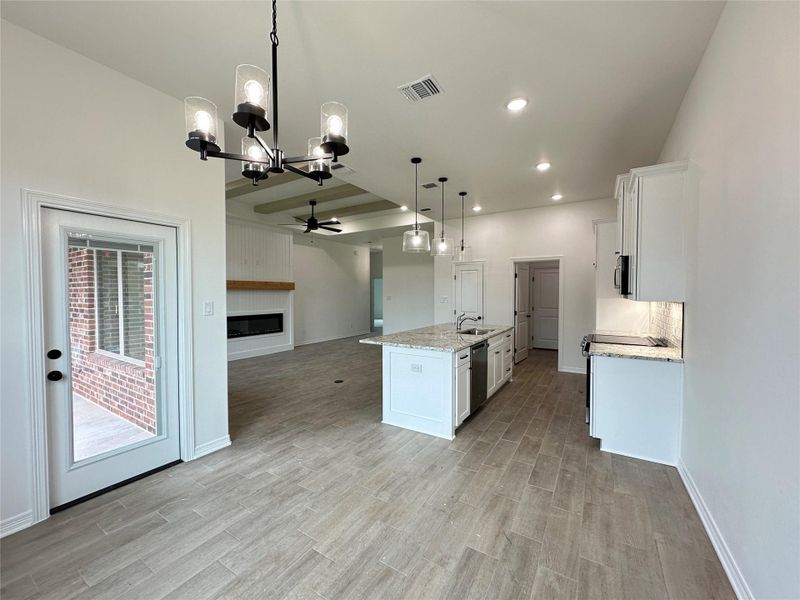 Open-concept living space featuring wood-finish flooring, a kitchen island with a stainless steel sink, recessed and pendant lighting, white shaker-style cabinetry, and a fireplace with a wood mantle