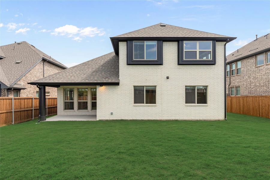 Two-story white brick home rear view with covered patio, large green lawn, and wood privacy fence