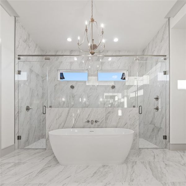 Freestanding soaking tub centered in a spacious bathroom featuring floor-to-ceiling marbled tile, dual glass-enclosed showers, and a chandelier