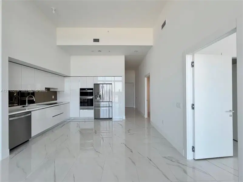 Contemporary kitchen featuring high-gloss white cabinetry, integrated stainless steel appliances, polished stone-finish tile flooring, and soaring ceilings