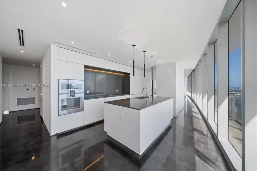 Modern kitchen featuring sleek flat-panel cabinetry, a central island with a solid-surface countertop, built-in stainless steel double ovens, floor-to-ceiling windows, and polished dark flooring Modern kitchen featuring sleek flat-panel cabinetry, a central island with a solid-surface countertop, built-in stainless steel double ovens, floor-to-ceiling windows, and polished dark flooring