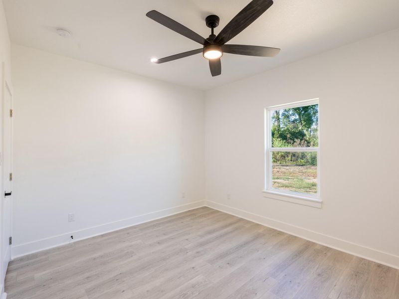This photo shows a bright, empty room with light wood flooring, white walls, and a ceiling fan. A window offers natural light and a view of greenery outside. Perfect for a bedroom or office space.