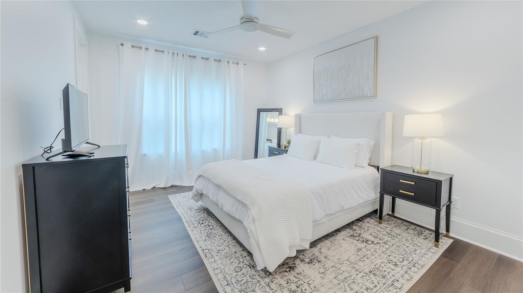 Bright and modern bedroom featuring a comfortable bed with white linens, nightstands with lamps, a wall-mounted TV, and a large window with sheer curtains. The room has sleek hardwood floors and a ceiling fan.