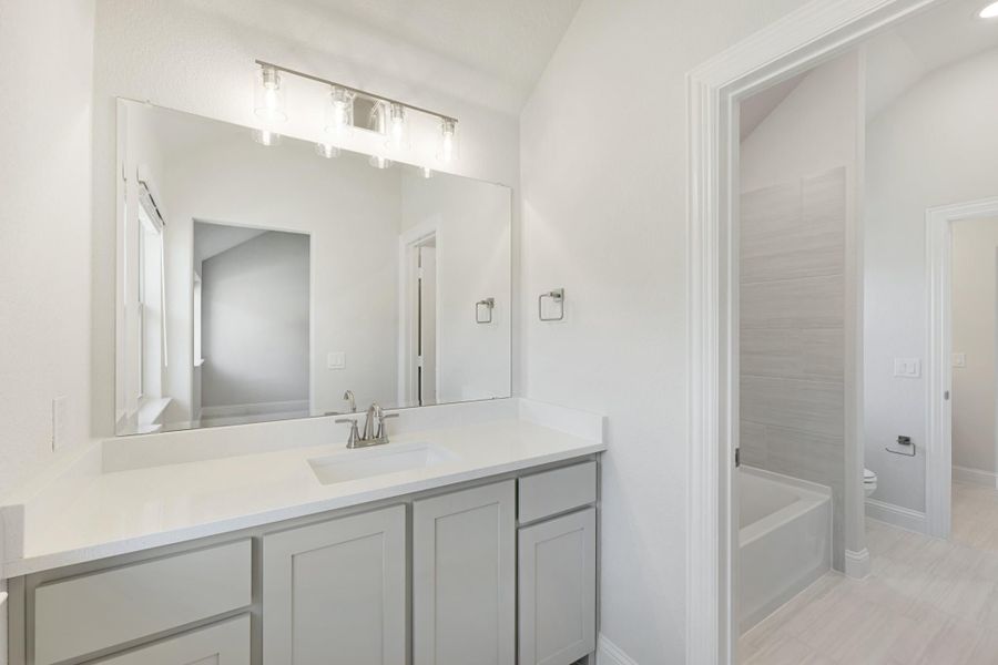 Bathroom with gray vanity, white countertop, large mirror, and tiled soaking tub visible through doorway