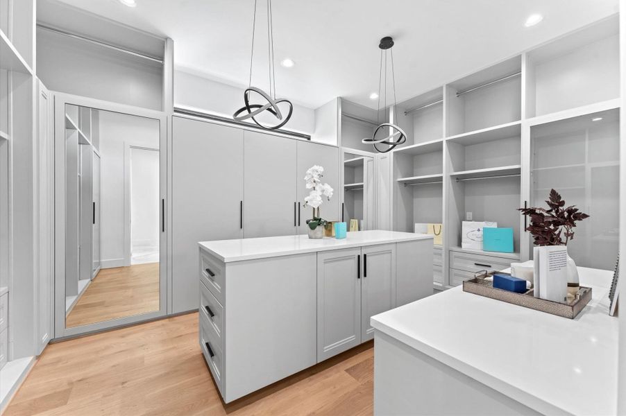 This is a spacious, modern walk-in closet featuring sleek white cabinetry, ample storage space, and a central island. It has contemporary pendant lighting and a large mirror, offering a bright and organized environment.