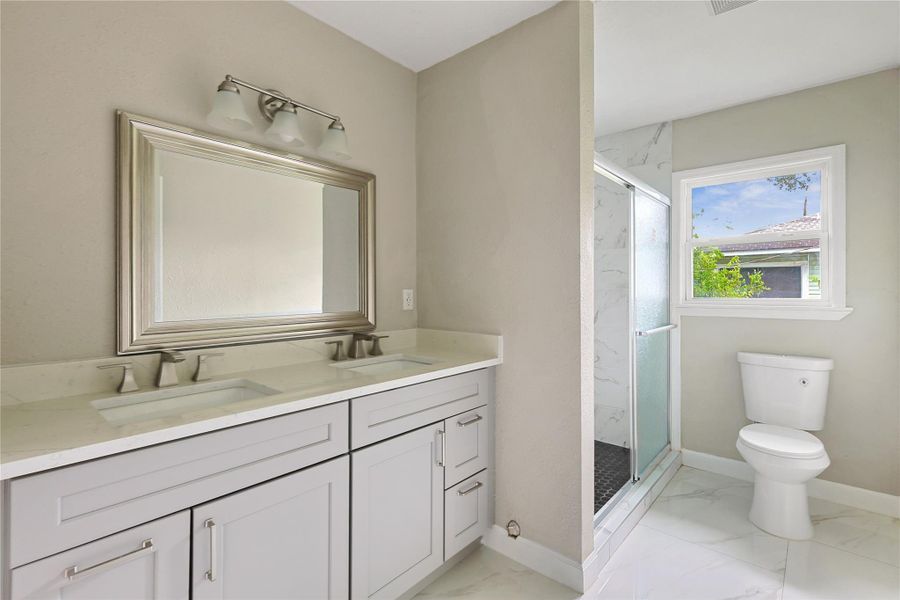 This bathroom features a modern double vanity with a large mirror, a spacious glass-enclosed shower, and a window providing natural light. The neutral tones and sleek fixtures create a clean, contemporary look.