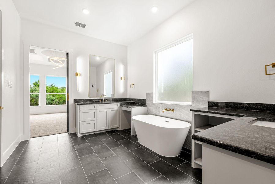 Sleek dark tile flooring leads to a spacious primary bathroom, complete with a freestanding soaking tub and dual vanities.