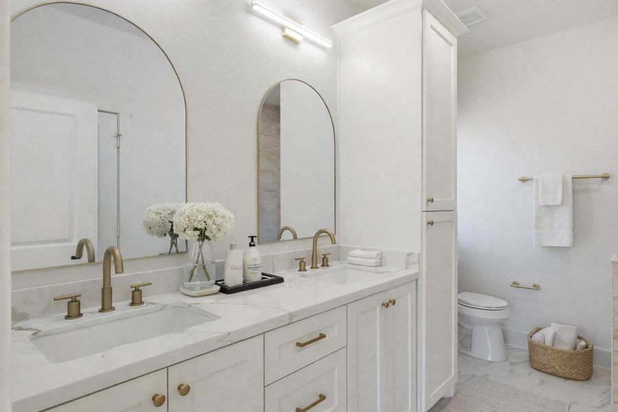 The ensuite bathroom features dual sinks with vanity lighting, custom cabinetry for extra storage, and a spacious walk-in shower. (staged)