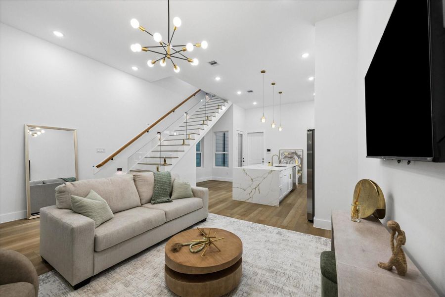 This modern living space features an open floor plan with elegant wood flooring and a staircase with glass railings. The room is well-lit with a contemporary chandelier and pendant lights over a sleek kitchen island. Neutral tones and minimalist decor create a sophisticated atmosphere. This modern living space features an open floor plan with elegant wood flooring and a staircase with glass railings. The room is well-lit with a contemporary chandelier and pendant lights over a sleek kitchen island. Neutral tones and minimalist decor create a sophisticated atmosphere.