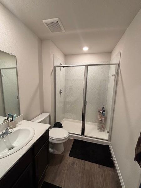 Modern bathroom featuring a glass-enclosed shower with white tiled walls, a dark wood-finish vanity with a white countertop and integrated sink, a commode, and wood-finish flooring
