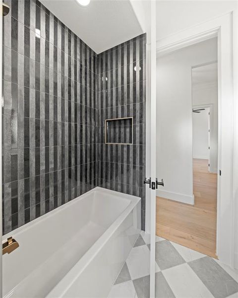 White soaking tub with gold-finish fixtures, set against a vertically-oriented striped tile shower surround featuring a recessed niche