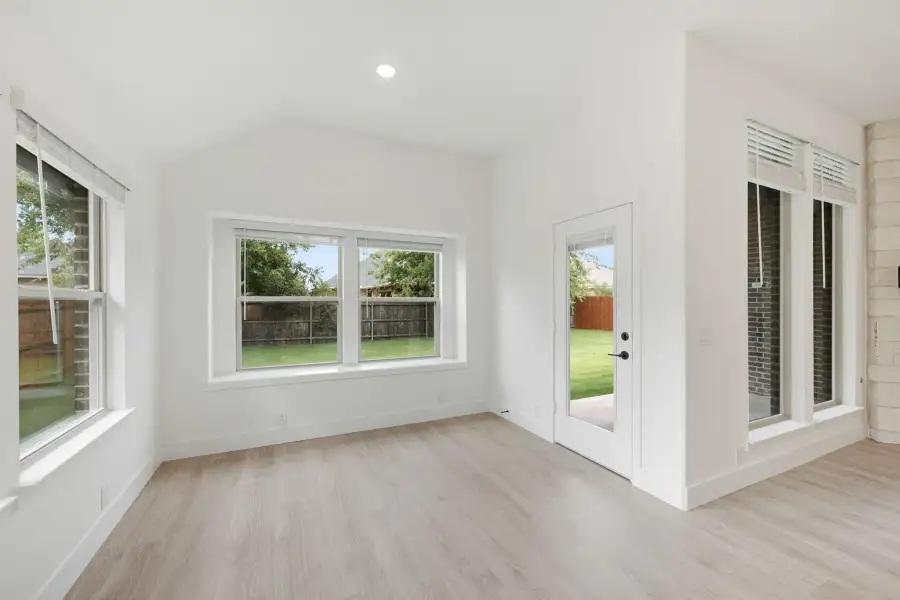 Empty living area with vaulted ceiling, light wood floors, large windows, and glass back door overlooking fenced yard